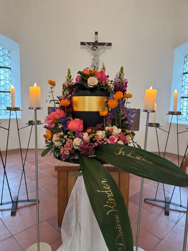 Schwarze Urne mit buntem Urnenkranz und Trauerschleife in einer Kapelle