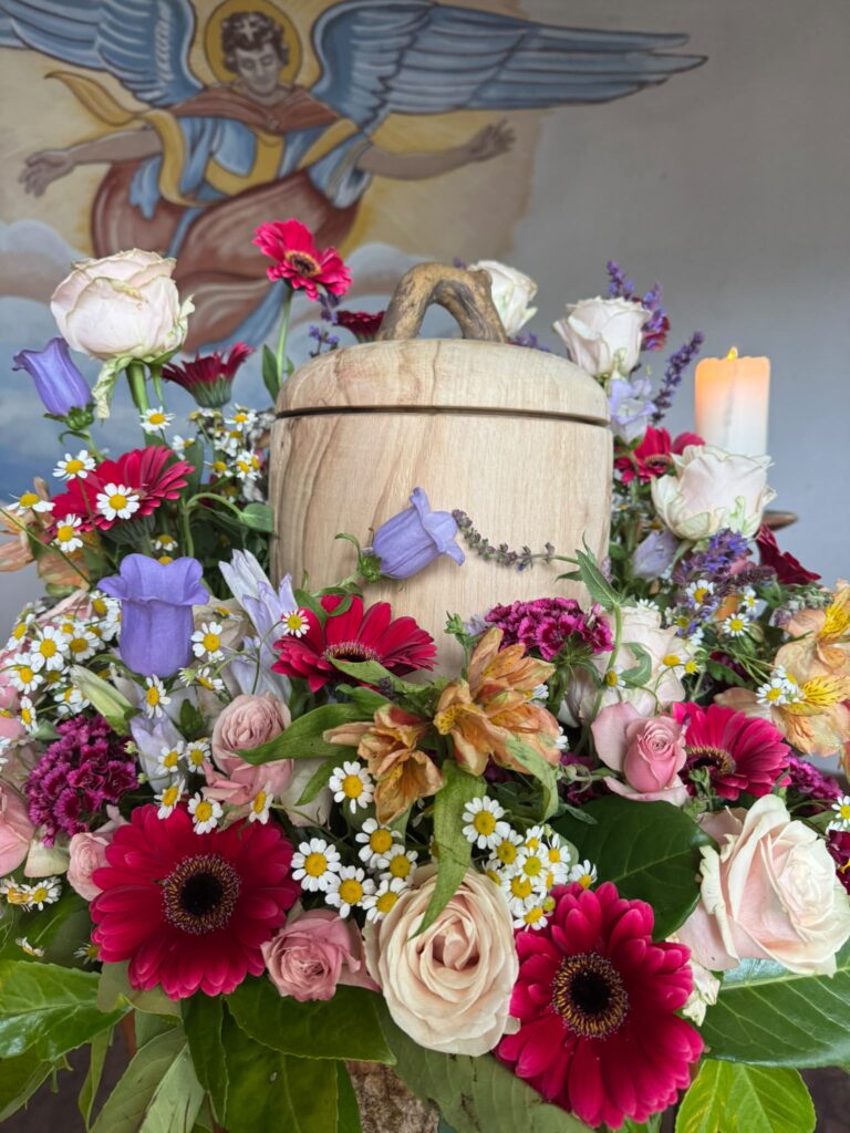 Holzurne umgeben von buntem Blumenschmuck mit Gerbera, Rosen und Glockenblumen