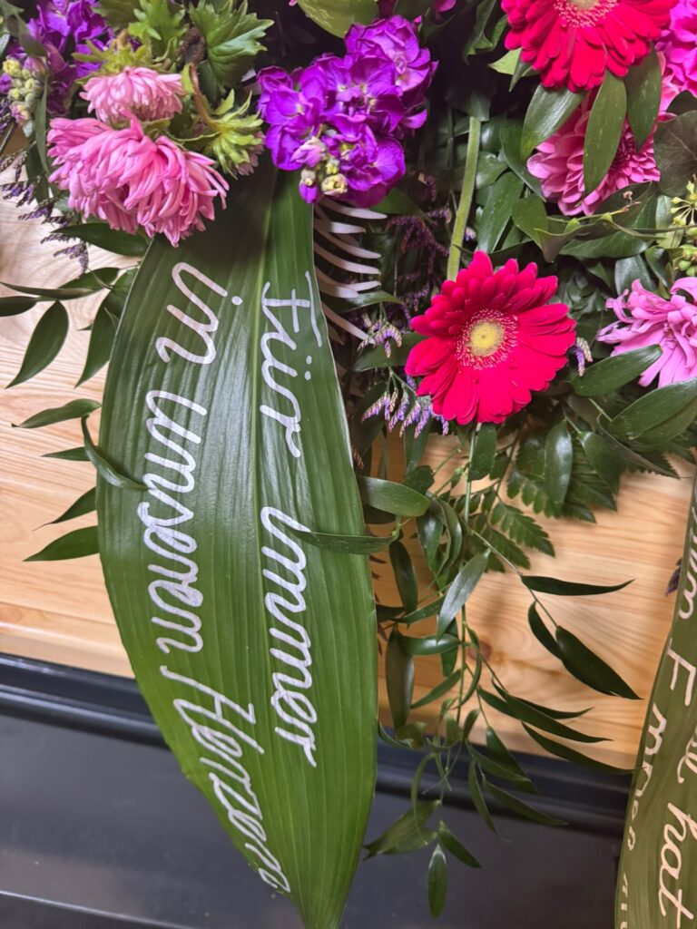 Sargschmuck mit pinken Blumen und grüner Trauerschleife mit Inschrift