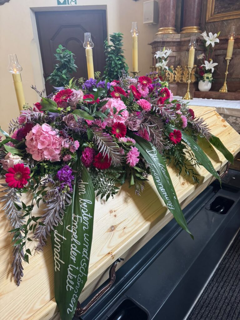 Holzsarg mit Sargbukett in Pink und Lila und Trauerschleife bei Kerzenlicht