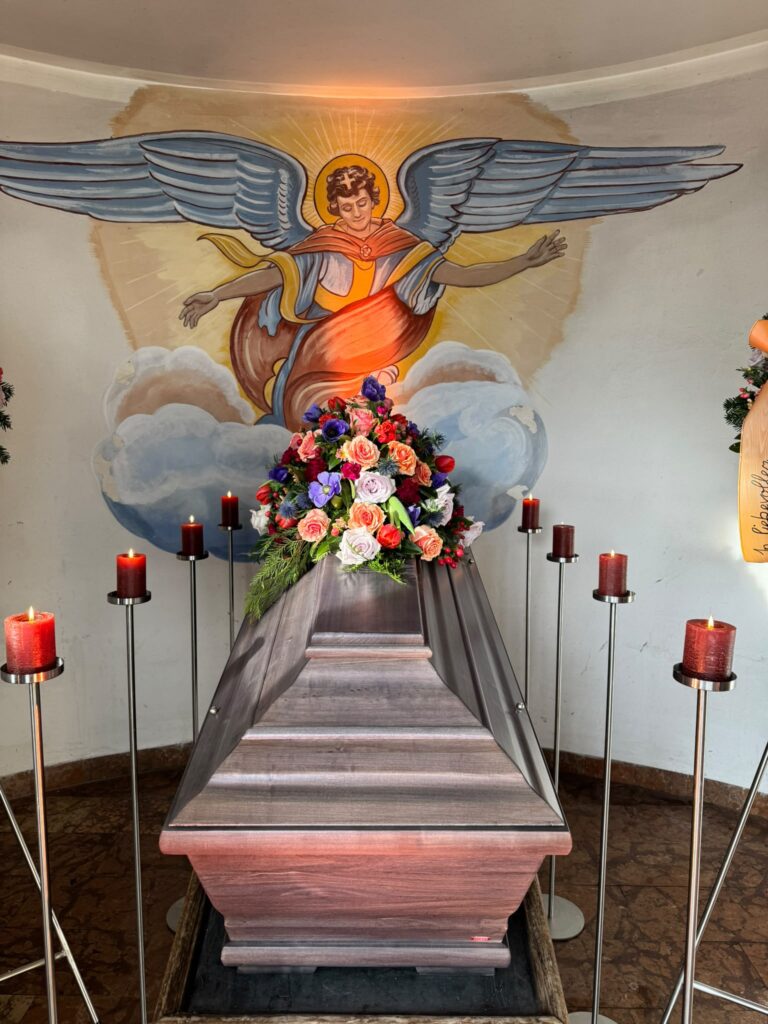 Bunter Blumenschmuck mit roten Kerzen vor einem Engel-Wandbild in einer Kapelle
